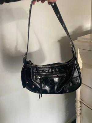 Svart axelväska i skinnlook - Snygg svart axelväska i glansig skinnimitation med silverfärgade nitar och dragkedjedetaljer. Väskan har en justerbar rem och en liten ficka framtill med dragkedja. Perfekt för dig som gillar en edgy och trendig stil.