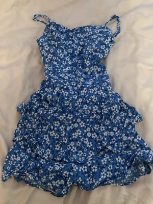 Blå blommig kortklänning med volang - Söt blå shortsklänning med vita blommor och volangdetaljer. Klänningen har smala axelband och en öppen rygg med knyt. Perfekt för sommarens alla tillfällen och ger en fräsch och ungdomlig look.