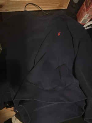 Mörkblå sweatshirt från Ralph Lauren - Klassisk mörkblå sweatshirt från Ralph Lauren med den ikoniska röda logotypen broderad på bröstet. Tröjan har rund halsringning och ribbade muddar vid ärmslut och nederkant. Perfekt för en avslappnad och stilren look. Nytt pris 2000, använt denna 3 gånger 