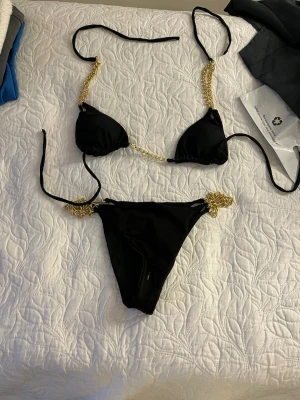 Svart bikini med guldkedjor - Snygg svart bikini med guldfärgade kedjedetaljer på både överdel och underdel. Klassisk trekantsmodell med knyt i sidorna och runt nacken. Perfekt för sommarens strandhäng eller poolparty.