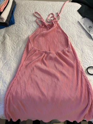 Rosa randig sommarklänning - Säljer en söt rosa klänning med tunna vita ränder och smala axelband. Klänningen har en avslappnad passform och är tillverkad i ett mjukt, lätt tyg som passar perfekt för varma dagar. Den har en kort längd och ett enkelt, stilrent uttryck.