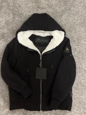 Svart hoodie Moose Knuckles bunny - Snygg svart hoodie från Moose Knuckles med dragkedja, vit fluffig foder i huvan och quiltat innerfoder. Har en ikonisk metall-logga på ärmen och praktiska fickor framtill. Perfekt för dig som vill ha både stil och komfort.