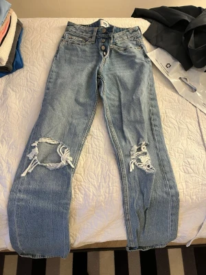 Ljusblå slitna jeans med hål - Säljer ett par ljusblå jeans med raka ben och stora slitningar vid båda knäna. Jeansen har hög midja, knappgylf och klassiska fem fickor. Perfekta för en avslappnad och trendig look.
