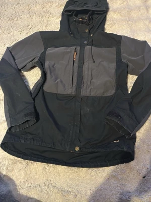 Svart och grå vindjacka med huva - Snygg vindjacka i svart och grått med justerbar huva och dragkedja framtill. Jackan har stora bröstfickor, kardborre vid ärmsluten och detaljer i läder på dragkedjan. Materialet är slitstarkt och passar perfekt för utomhusaktiviteter.