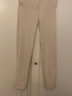 Beige linnebyxor med rak passform - Snygga beige linnebyxor med rak passform och hög midja. Byxorna har en enkel design utan synliga fickor eller detaljer, vilket ger en clean och stilren look. Perfekta för dig som gillar minimalistisk stil och vill ha något luftigt och bekvämt i garderoben.