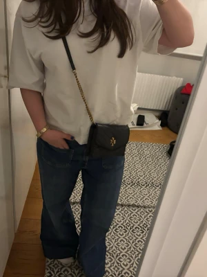 Svart axelväska från Tory Burch - Snygg svart axelväska från Tory Burch med guldfärgad kedjerem och logga framtill. Väskan har en kompakt och stilren design som passar perfekt till jeans eller klänning. Den är helt perfekt i storlek 