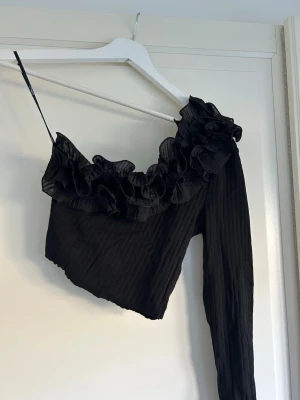 Svart oneshoulder topp med volang - Trendig svart oneshoulder topp med volangdetaljer längs halsringningen. Toppen har ribbad struktur!!🖤