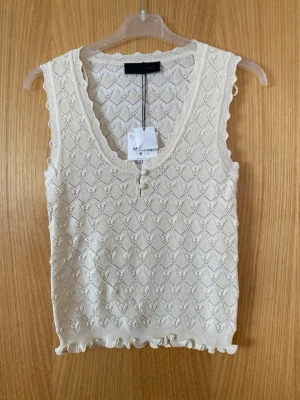 Vit stickad linnetopp från Zara  - Säljer en vit stickad linnetopp från Zara med v-ringning och dekorativa volangdetaljer vid ärm och nederkant. Toppen har ett diskret mönster och är ärmlös, perfekt för att skapa en söt och trendig look.