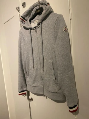 Grå hoodie från Moncler med dragkedja - Dem som tror den e fejk för de står storlek S så har moncler bara skrivit med nummer på jackor och västar!!!     Snygg grå hoodie från Moncler med hel dragkedja, huva med dragsko och fickor framtill. Detaljer med röd, vit och marinblå rand på muddar och huva samt Moncler-logga på ärmen. Tillverkad i mjuk bomullsblandning för extra komfort.