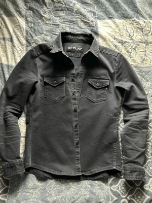 Mörkgrå overshirt från Replay - Snygg svart jeansjacka från Replay med klassisk krage, två bröstfickor med lock och knapp samt knäppning framtill. Jackan har raka ärmar och en stilren, tidlös design som passar till det mesta. Perfekt för dig som gillar en clean och enkel look. 9/10 skick,  nypris 2599.