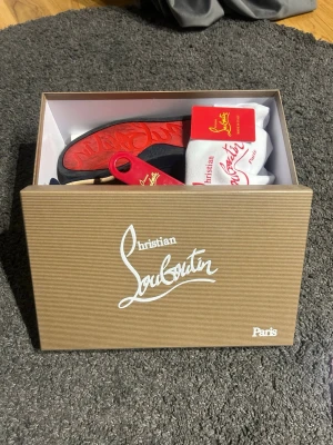 Christian Louboutin sneakers navy/röd - Snygga sneakers från Christian Louboutin i marinblå mocka med ikonisk röd sula. Skorna har lågt skaft och klassisk snörning. Kommer i originalkartong med dustbag och skohorn. Perfekta för dig som vill sticka ut med exklusiv stil.