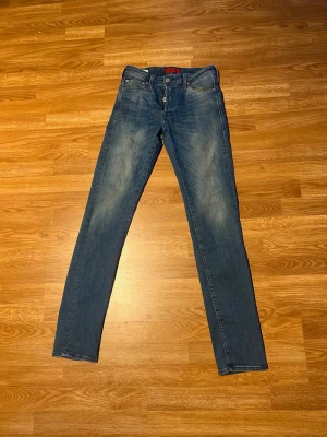 Blå slim fit jeans från Jack and Jones  - Snygga blå slim fit jeans från Jack and Jones med klassisk femficksdesign och slitna detaljer på låren. Jeansen har knappgylf och är tillverkade i ett mjukt denimtyg som sitter skönt. Perfekta för en avslappnad och trendig look.