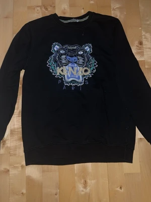 Svart sweatshirt från Kenzo - Cool svart sweatshirt från Kenzo med ikoniskt tigerbroderi och Kenzo Paris-text på bröstet. Tröjan har rund halsringning och ribbade muddar. 