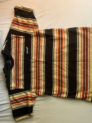 Randig t-shirt från Levi's - Säljer en snygg randig t-shirt från Levi's med broderad logga på bröstet. T-shirten har breda ränder i svart, beige, orange och vitt. Klassisk passform med korta ärmar och rund hals. Perfekt för dig som gillar retrovibbar och streetstyle.