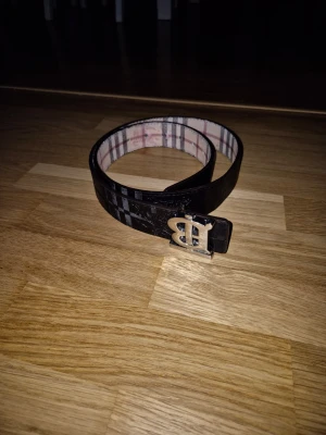 Svart och rutig burberry bälte - Snyggt skärp med svart utsida och klassiskt rutigt mönster på insidan. Skärpet har ett metallspänne med BB-logga och passar perfekt för att lyfta din outfit. Stilsäker accessoar med trendig design.