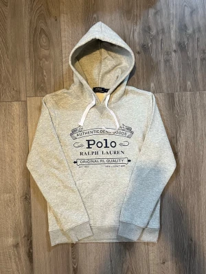 Grå hoodie från Polo Ralph Lauren - Snygg grå hoodie från Polo Ralph Lauren med stor mörkblå logga och text på bröstet🤗 använd ett par gånger men inga fel på den🙌🏻 inget äkthetsbevis tyvärr. Hör av er vid snabbaffär samt funderingar🤩