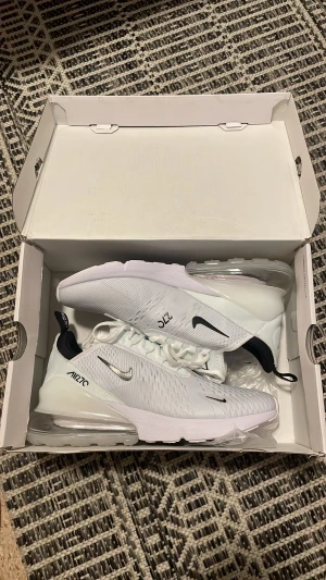 Nike Air Max 270 vita sneakers - Nike Air Max 270 sneakers i vitt med svarta detaljer och synlig Air Max-sula. Ovandelen är i mesh för bra ventilation och har klassisk Nike-logga på sidan. Skorna har vita snören och en chunky, genomskinlig sula som ger en sportig vibe.
