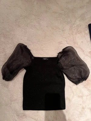 Svart topp med puffärmar från Gina Tricot - Trendig svart topp från Gina Tricot med glittrig finish och stora transparenta puffärmar. Toppen har en tight passform i kroppen och fyrkantig ringning. Perfekt för dig som vill sticka ut med en cool och elegant look.