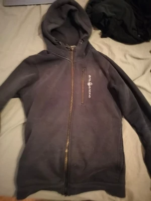 Svart hoodie från Peak Performance - Snygg svart hoodie från Peak Performance med dragkedja framtill och logga på bröstet. Klassisk huva och mjukt material som känns skönt mot huden. Perfekt för en avslappnad och sportig stil.