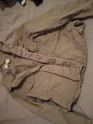 Grön jeansjacka med stora knappar - Snygg grön jeansjacka med stora knappar framtill och två bröstfickor. Jackan har en avslappnad passform och rå kant nedtill för en trendig look. Perfekt för dig som vill ha en cool och enkel stil.