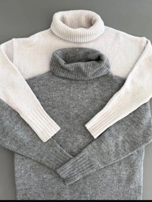 Två polotröjor i stickat material - Säljer två stickade polotröjor, en i ljusbeige och en i grått. Båda har ribbstickad krage och muddar, samt lång ärm. Perfekta för kalla dagar och supermysiga att bära. Materialet ser mjukt och värmande ut.