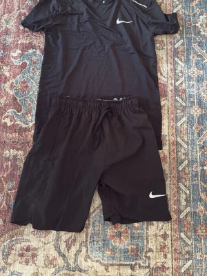 Nike miler t shirt short set - Säljer ett svart träningsset från Nike med t-shirt och shorts. Båda plaggen har den klassiska Nike-loggan i vitt. T-shirten har rund hals och korta ärmar, shortsen har resårmidja med dragsko för skön passform. Perfekt för gym, löpning eller annan träning.