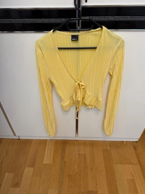 Gul ribbad omlottopp med knyt - Supersöt ljusgul omlottopp från Gina Tricot med ribbad struktur och knyt framtill. Toppen har långa ärmar, volangkant nedtill och v-ringad urringning. Perfekt till sommaren eller våren.