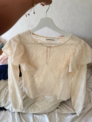 Vit spetsblus från Carin Wester - Säljer en vit spetsblus från Carin Wester med blommigt mönster och volangdetaljer på axlarna. Blusen har rund halsringning och en liten knapp framtill. Långärmad och tillverkad i ett tunt, transparent material som känns riktigt fräscht.💕