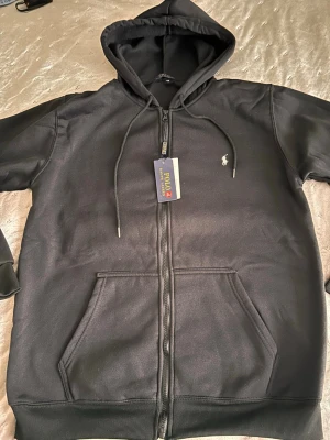 Svart hoodie från Polo Ralph Lauren - Svart hoodie med dragkedja från Polo Ralph Lauren. Klassisk design med huva och snören, två stora fickor framtill och ikonisk broderad logotyp på bröstet. Materialet är mjukt och bekvämt, perfekt för en avslappnad look.