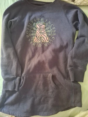 Mörkblå hoodie med zebramönster - Mysig mörkblå hoodie med stor ficka framtill och ett stort A i zebramönster på bröstet. Runt bokstaven står det ' GIRL PRINCESS ROYALTY CLUB' i grönt. Långa ärmar och mjukt material, perfekt för chill dagar.