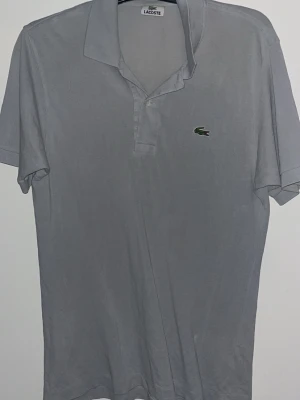 Grå pikétröja från Lacoste - Klassisk grå pikétröja från Lacoste med korta ärmar och den ikoniska gröna krokodilloggan på bröstet. Tröjan har krage och knäppning framtill. Perfekt till en avslappnad men ändå stilren look.