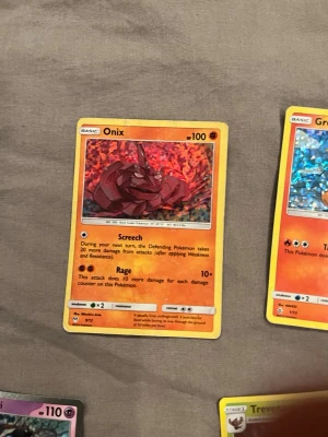 Onix Pokémonkort holografisk - Säljer ett holografiskt Onix Pokémonkort med orange ram och färgstark bakgrund. Kortet har attacker som Screech och Rage och visar Onix i en röd och orange miljö. Perfekt för samlare eller Pokémonfans.