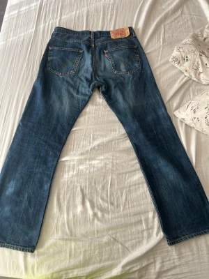 Levi's blå raka jeans - Säljer ett par klassiska blå jeans från Levi's med rak passform och fem fickor. Jeansen har en snygg tvätt och den ikoniska läderlappen bak i midjan. Perfekta för dig som gillar tidlös stil och vill ha ett par jeans som funkar till allt. De har en liten fläck på bakfickan som knappt syns
