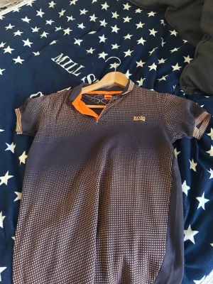Mörkblå prickig polotopp Hugo Boss - Snygg mörkblå polotopp från Hugo Boss med orange detaljer och små vita prickar över hela plagget. Kortärmad modell med klassisk krage och logga broderad på bröstet. Perfekt för dig som gillar stilrena men ändå unika toppar.