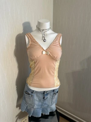 Beige linnetopp med spetsdetaljer - Söt beige linnetopp med spets i sidorna och en liten rosett framtill. Toppen har v-ringning och är ärmlös, med figurnära passform. Perfekt för sommarens alla outfits och ger en romantisk vibe.