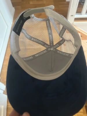 Blå truckerkeps från Maggiore - Snygg blå truckerkeps från Maggiore med mesh på sidorna och justerbar snapback. Framsidan är i mjukt tyg och har en metallbricka med Maggiore-loggan. Perfekt för en avslappnad streetwear-look.