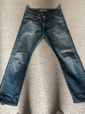 Blå raka jeans från Levi's - Säljer ett par klassiska blå jeans från Levi's med rak passform och slitningar för en cool vintage-look. Jeansen har fem fickor, knappgylf och den ikoniska röda Levi's-lappen bak. Tillverkade i slitstark denim som passar perfekt till en avslappnad stil.