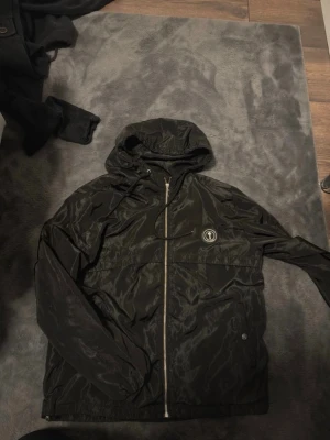 Svart vindjacka med huva från Galagowear - Säljer en svart vindjacka från Galagowear med huva och dragkedja framtill. Jackan har ett stilrent tryckt logomärke på bröstet och är tillverkad i ett glansigt, lätt material som passar perfekt för blåsiga dagar. Justerbar huva med snören och två sidofickor.