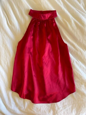 Röd halterneck topp  - Snygg röd halterneck topp i glansigt satintyg. Toppen har hög krage och är ärmlös med en lös passform. Perfekt för dig som vill sticka ut med en elegant och trendig look.