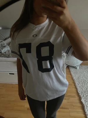 Vit oversized t-shirt med tryck 87 - Säljer en vit oversized t-shirt med svart tryck av siffran 87 på framsidan och sportiga ränder på ärmarna. T-shirten har v-ringad hals och är kortärmad. Perfekt för en avslappnad och trendig look.