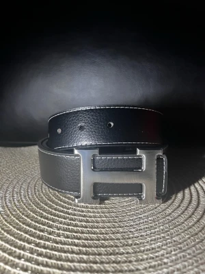 Svart och blått bälte - Snyggt blått och avart bälte i läder med vit kontrastsöm och ett stort, silverfärgat metallspänne. Perfekt accessoar för att lyfta din outfit med en stilren och klassisk touch.