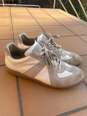 Maison Margiela vita och beige sneakers - Maison Margiela sneakers i vitt och beige med klassisk låg silhuett. Ovandelen är i en mix av mocka och slätt läder, med gummisula och snörning. Ikonisk design med diskreta detaljer och Maison Margiela-logga under sulan.