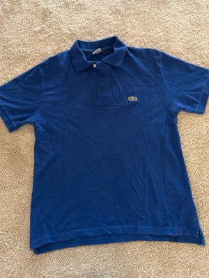 Blå pikétröja från Lacoste - Klassisk blå pikétröja från Lacoste med krage och knappar framtill. Tröjan har den ikoniska krokodilloggan på bröstet och är tillverkad i mjuk bomull. Perfekt till en avslappnad och stilren look. Pris kan diskuteras 