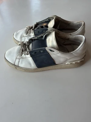 Valentino open vita  - Säljer ett par vita Valentino Garavani sneakers med svart band över vristen och nitar på hälen. Skorna är låga och har en chunky sula. Tillverkade i läder med klassisk snörning och coola detaljer som ger en lyxig vibe.