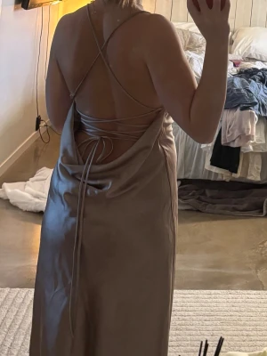 Champagnefärgad långklänning med snörning - Säljer en elegant långklänning i champagnefärgad satin med öppen rygg och tunna axelband som korsas och knyts i ryggen. Klänningen har en mjuk, glansig finish och en smickrande siluett som faller snyggt längs kroppen. Klänningen är ankel lång