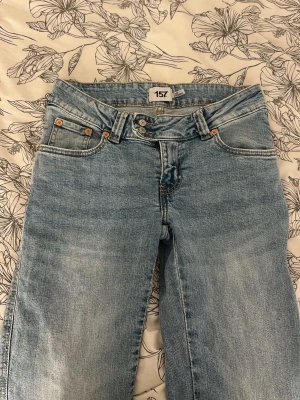 Ljusblå raka jeans från 157 - Säljer ett par korta ljusblå flare jeans från 157 med klassisk femficksmodell. Jeansen har dubbla knappar i midjan och detaljer i kopparfärg vid fickorna. Perfekta för en avslappnad och trendig look.