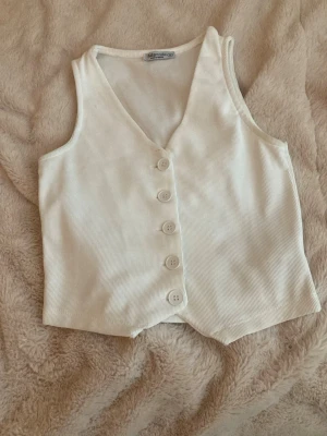 Vit väst från fullamoda med knappar - Stilren vit väst från fullamoda i ribbad bomull. Västen har v-ringning och fem stora knappar framtill. Perfekt att styla över en skjorta eller t-shirt för en trendig look.