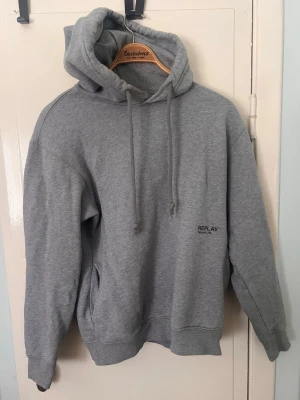 Grå hoodie från Replay - Säljer en stilren grå hoodie från Replay med trycket 'Replay Second Life' på bröstet. Hoodien har huva med dragsko och en klassisk, avslappnad passform. Perfekt för dig som gillar enkel och clean streetwear. Materialet är mjukt och skönt, troligen bomullsblandning.