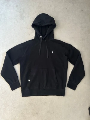 Svart hoodie från Polo Ralph Lauren - Klassisk svart hoodie från Polo Ralph Lauren med vit broderad logga på bröstet. Hoodien har huva med dragsko, stor magficka och ribbade muddar vid ärmar och nederkant. Perfekt för en avslappnad och stilren look. Hoodien är i 9/10 i skick. Den är i storlek M men passar även S!