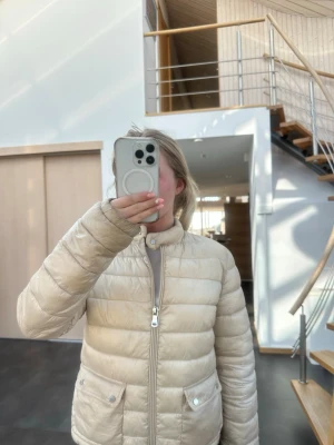 Beige pufferjacka med dragkedja - Snygg beige pufferjacka med hög krage och dragkedja framtill. Jackan har två framfickor med knappstängning och quiltad design. Perfekt för kalla dagar och ger en stilren look.